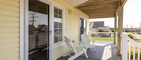 A covered porch with a wooden railing, a sliding glass door, and two Adirondack chairs.