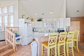 A kitchen with white cabinets, a built-in microwave, a sink, and a dining area with yellow chairs. - Ocean Escape: Oceanside, pool, hot tub, pool table, easy beach access (Kill Devil Hills)
