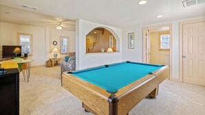 Une table de billard dans une pièce dotée d’un ventilateur de plafond, d’un téléviseur et d’une fenêtre avec des stores.