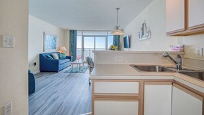 A modern kitchen with a central island, a dining area with a blue sofa, and a view of the outdoors through large windows. - Oceanfront 1BD-Baywatch Resort-Family Friendly (North Myrtle Beach)