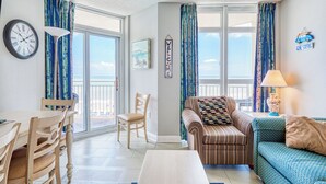 Un salon comprenant une table à manger, des chaises, un canapé et une lampe. Il y a une horloge au mur et une pancarte sur laquelle on peut lire « Bienvenue ».