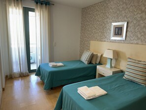 A hotel room with two beds, a nightstand with a lamp, a framed picture on the wall, and a view of the outside through a window with curtains.