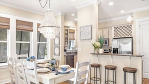A dining area with a white table set for four, surrounded by white chairs. A chandelier hangs above, and a kitchen with stainless steel appliances is visible in the background.