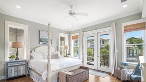 A bedroom with a four-poster bed, a ceiling fan, a large window with a view of the outdoors, and a wicker bench.