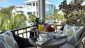 A wicker table set with breakfast items, including croissants, strawberries, and orange juice, on a balcony with a view of buildings and palm trees.