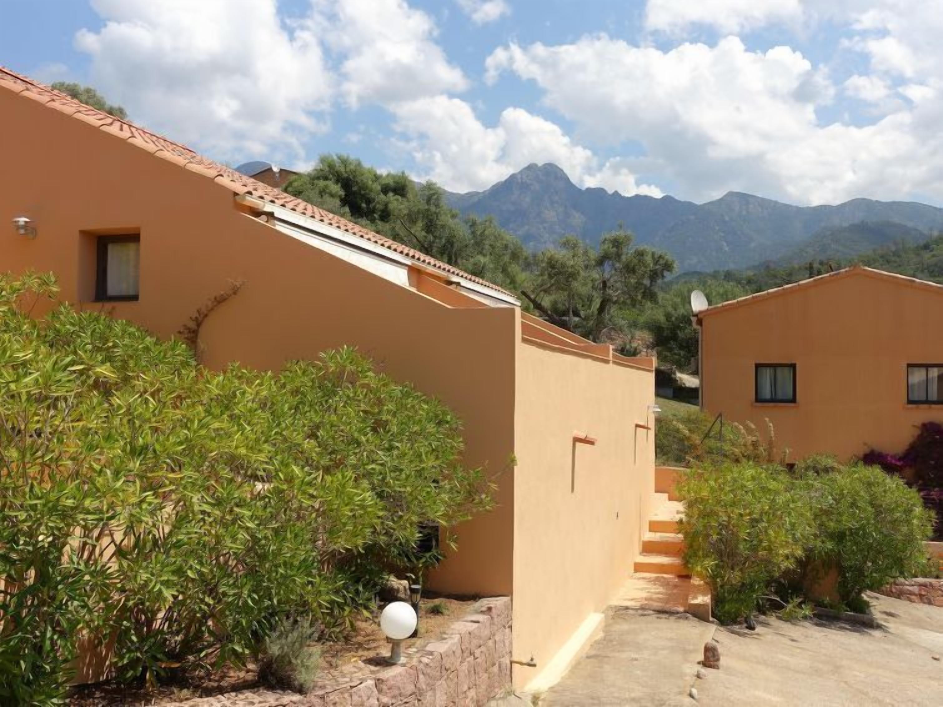 Un bâtiment doté d’un toit plat et d’une petite fenêtre, entouré de verdure et avec des montagnes en arrière-plan.