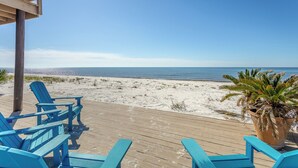 Une terrasse en bois avec des fauteuils Adirondack bleus, donnant sur une plage de sable et l’océan.