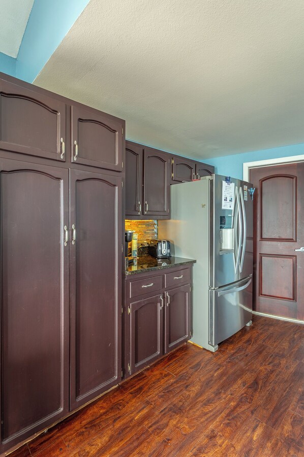 A kitchen with dark wooden cabinets, a stainless steel refrigerator, and a wooden floor. - BEACH FRONT gathering spot for family, friends and pups. WINTER RATES AVAILABLE! (Alligator Point)