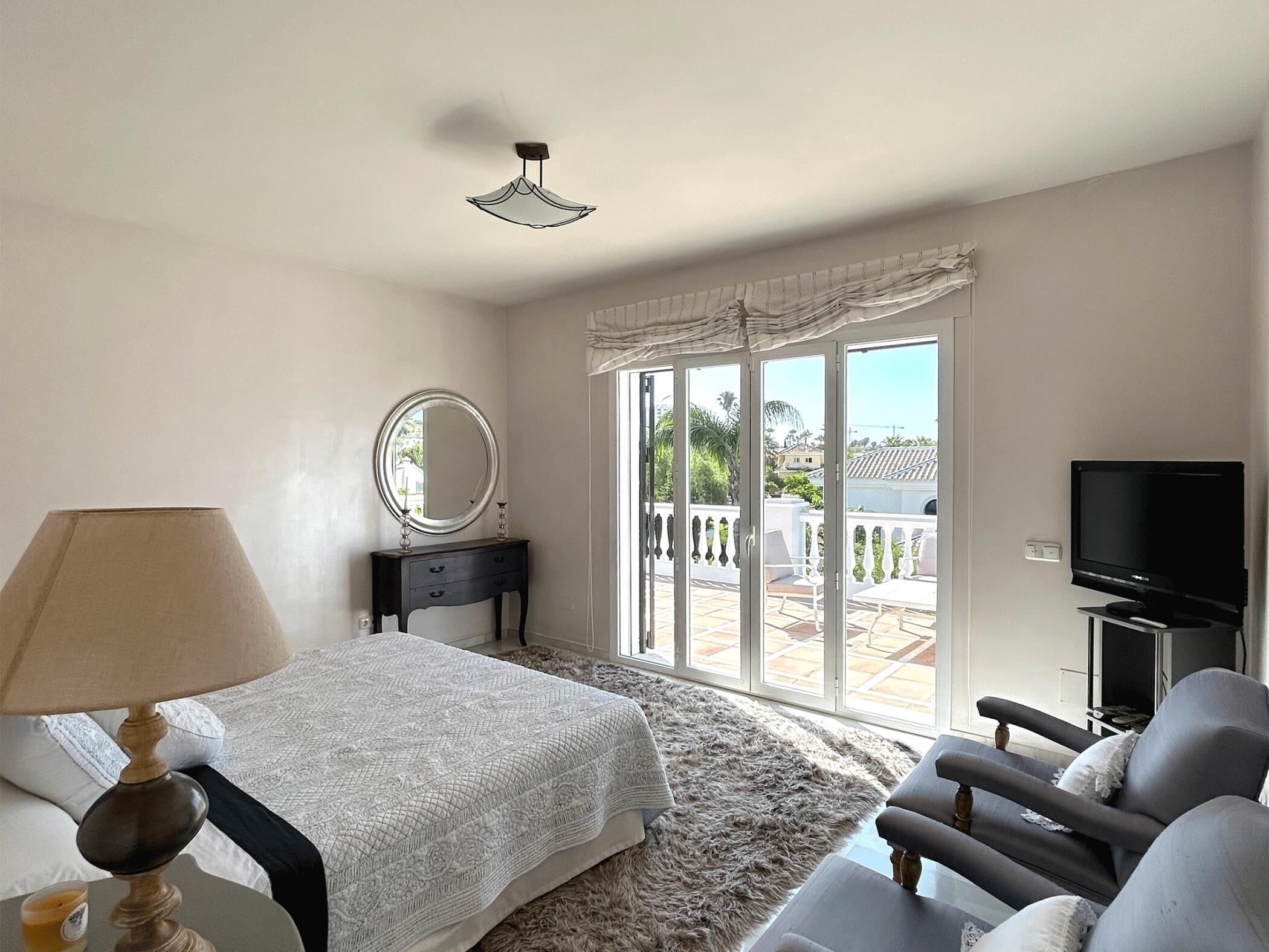 Une chambre à coucher comprenant un lit, une table de chevet avec une lampe, un miroir, une télévision et une vue sur le balcon extérieur.