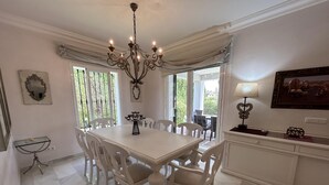 Une salle à manger avec une table et des chaises blanches, un lustre et une vue dégagée sur l’extérieur grâce à de grandes fenêtres.