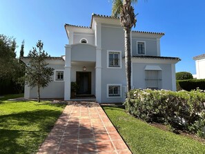 A two-story house with a paved driveway leading to a front door, surrounded by well-maintained grass and a palm tree. - Villa Maisa Luxury Marbella by Málaga Planners (Marbella)