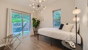 A modern bedroom with a large bed, a ceiling light fixture, a floor lamp, and a view of greenery through the windows. - Nashville Charmer (Nashville)