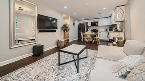 A modern living room with a sofa, a coffee table, and a TV. There is a kitchen area with cabinets and appliances in the background. - Nashville Charmer (Nashville)