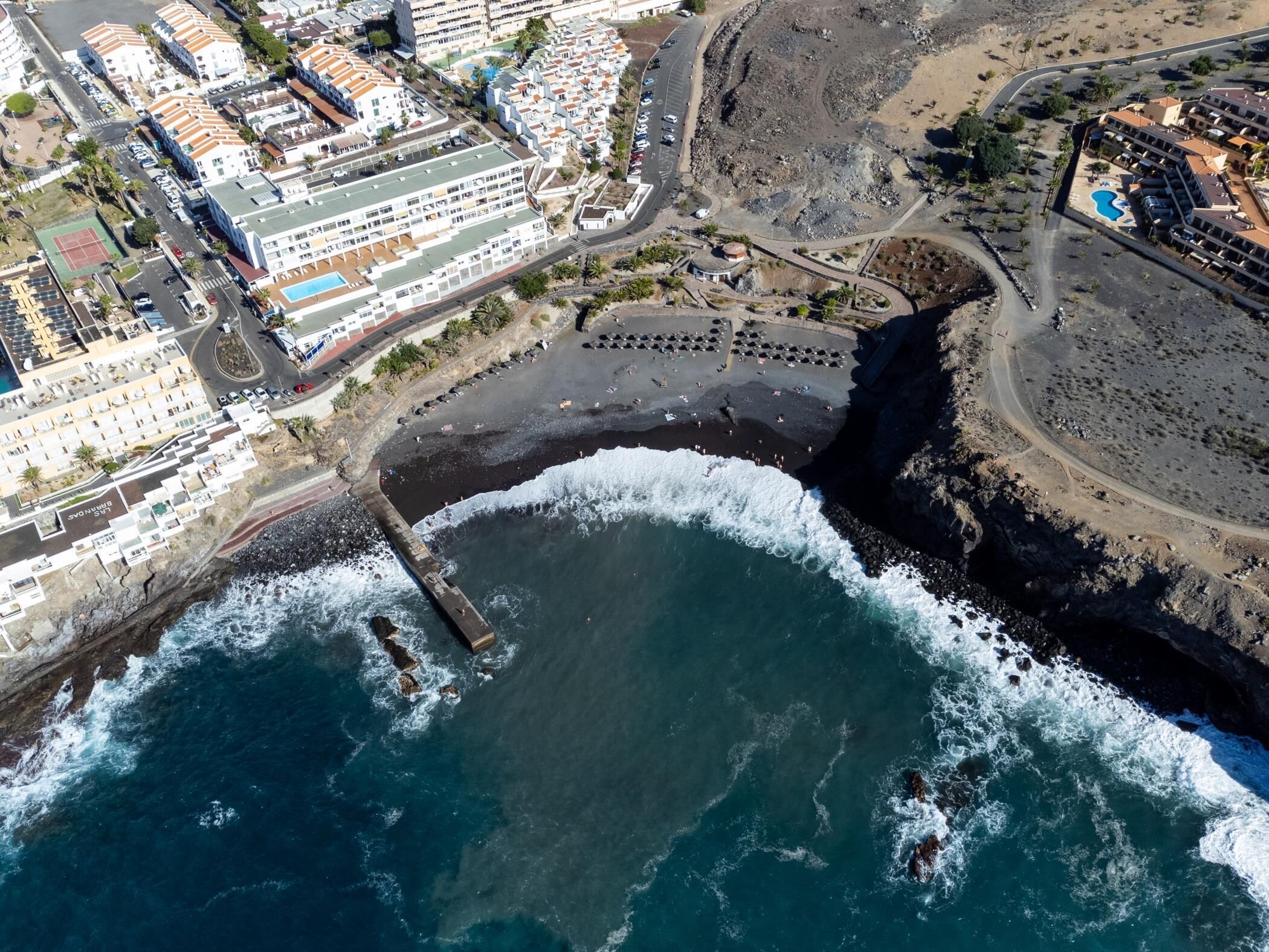 An aerial view of a coastal area with a beach, a rocky shoreline, and a hotel complex with multiple buildings and a swimming pool.