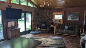 A cozy living room with a stone fireplace, a sofa, a television, and a kitchen area with wooden cabinets and a large window.