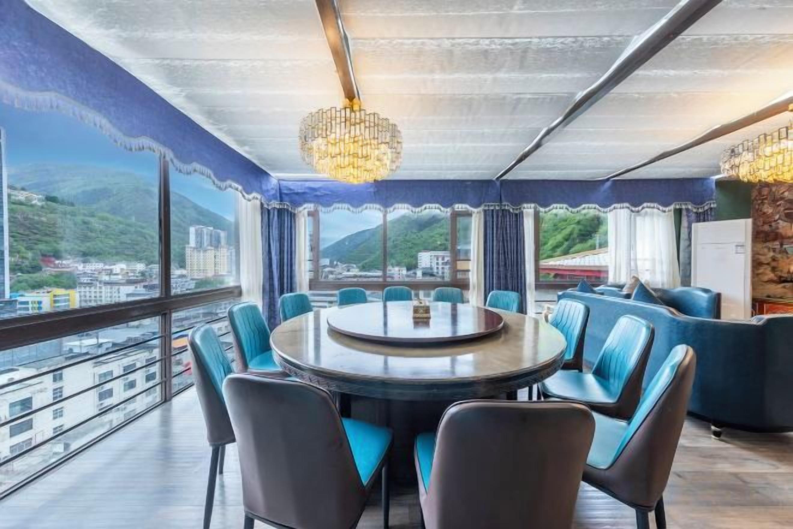 Uma área de jantar com mesa redonda e cadeiras, amplas janelas com vista para edifícios e montanhas, e um candelabro.