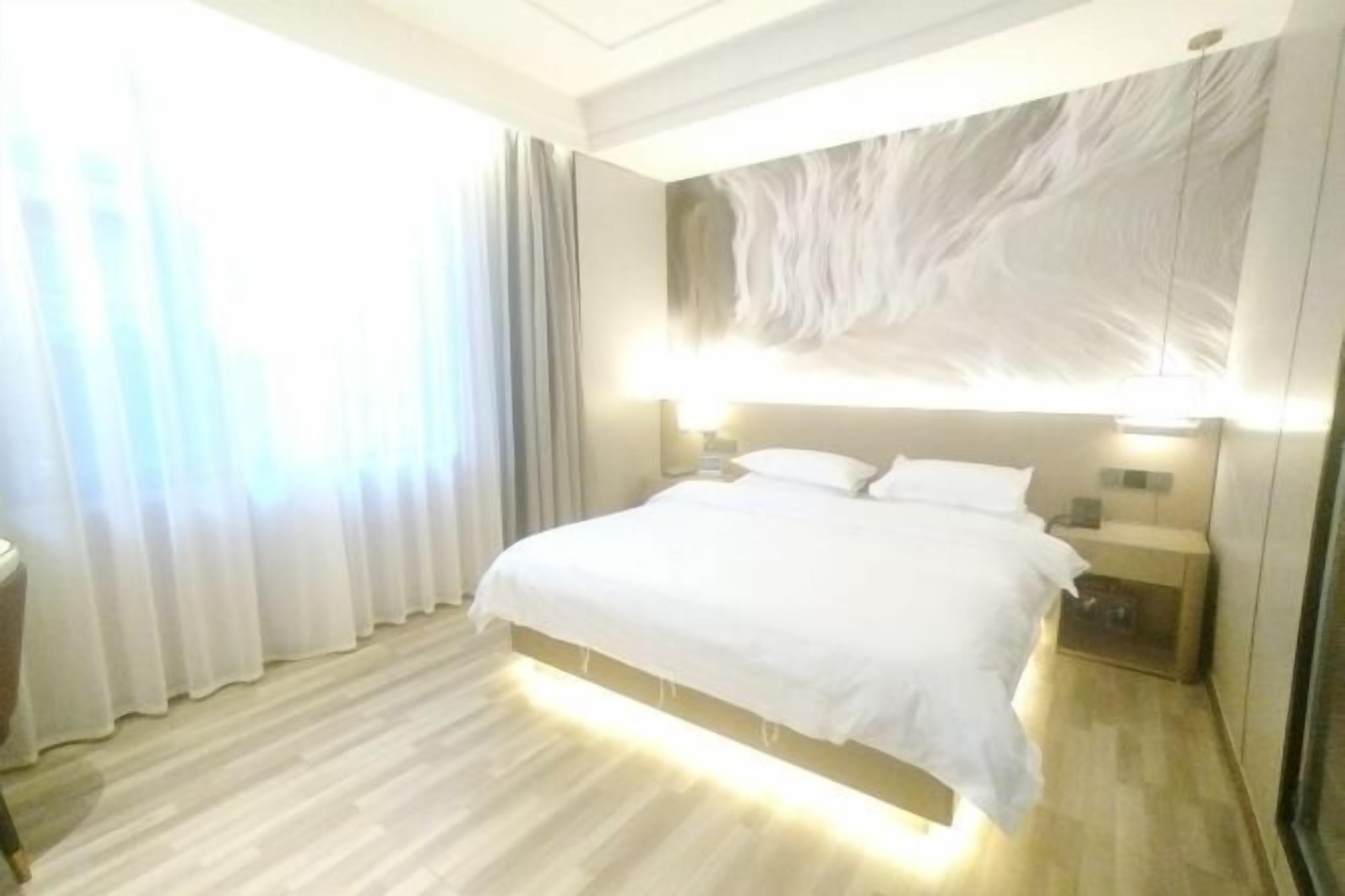 Quarto de hotel com uma cama grande, mesas de cabeceira, luminária na parede e uma janela com cortinas.