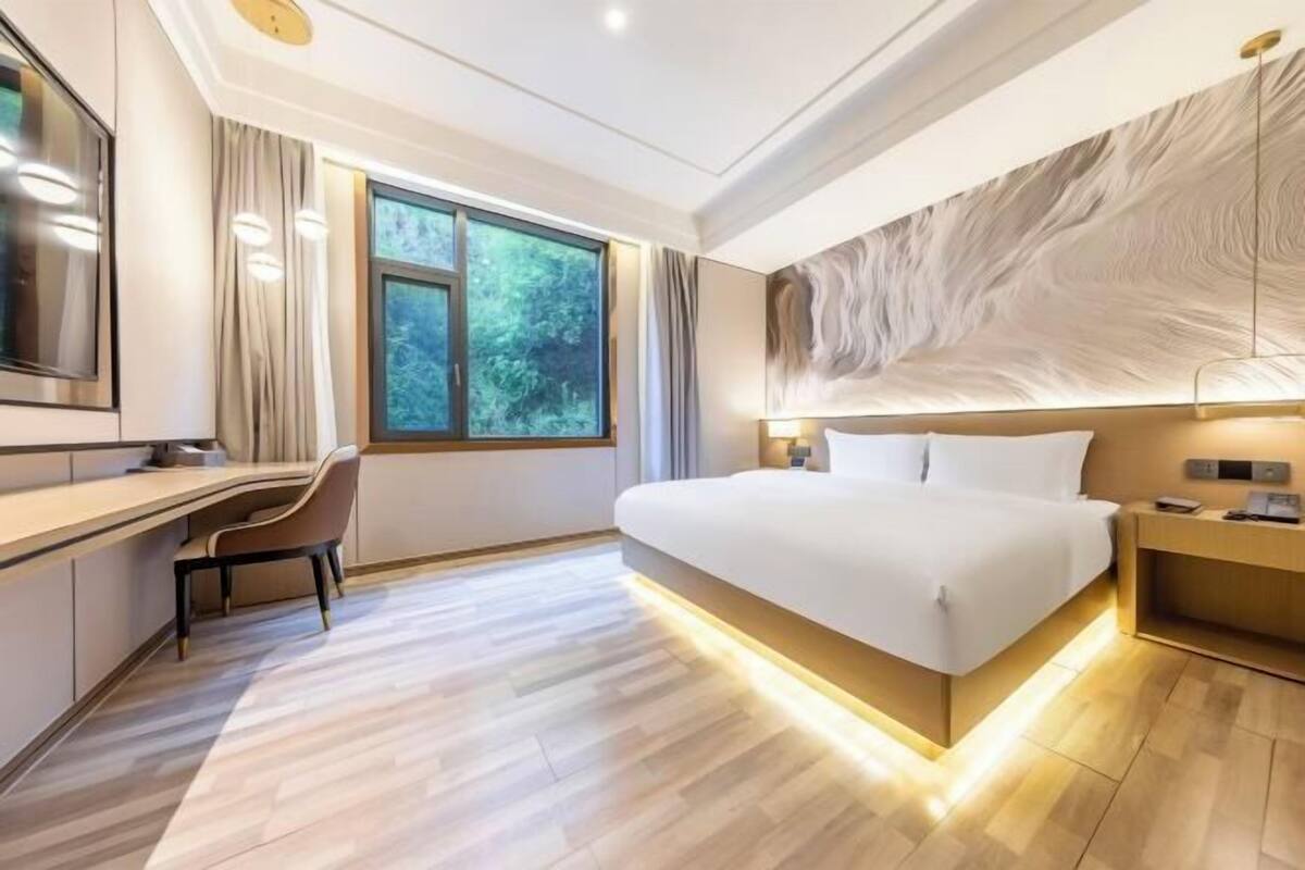 A modern hotel room with a large bed, a desk, and a view of greenery through the window.