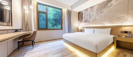 A modern hotel room with a large bed, a desk, and a view of greenery through the window.