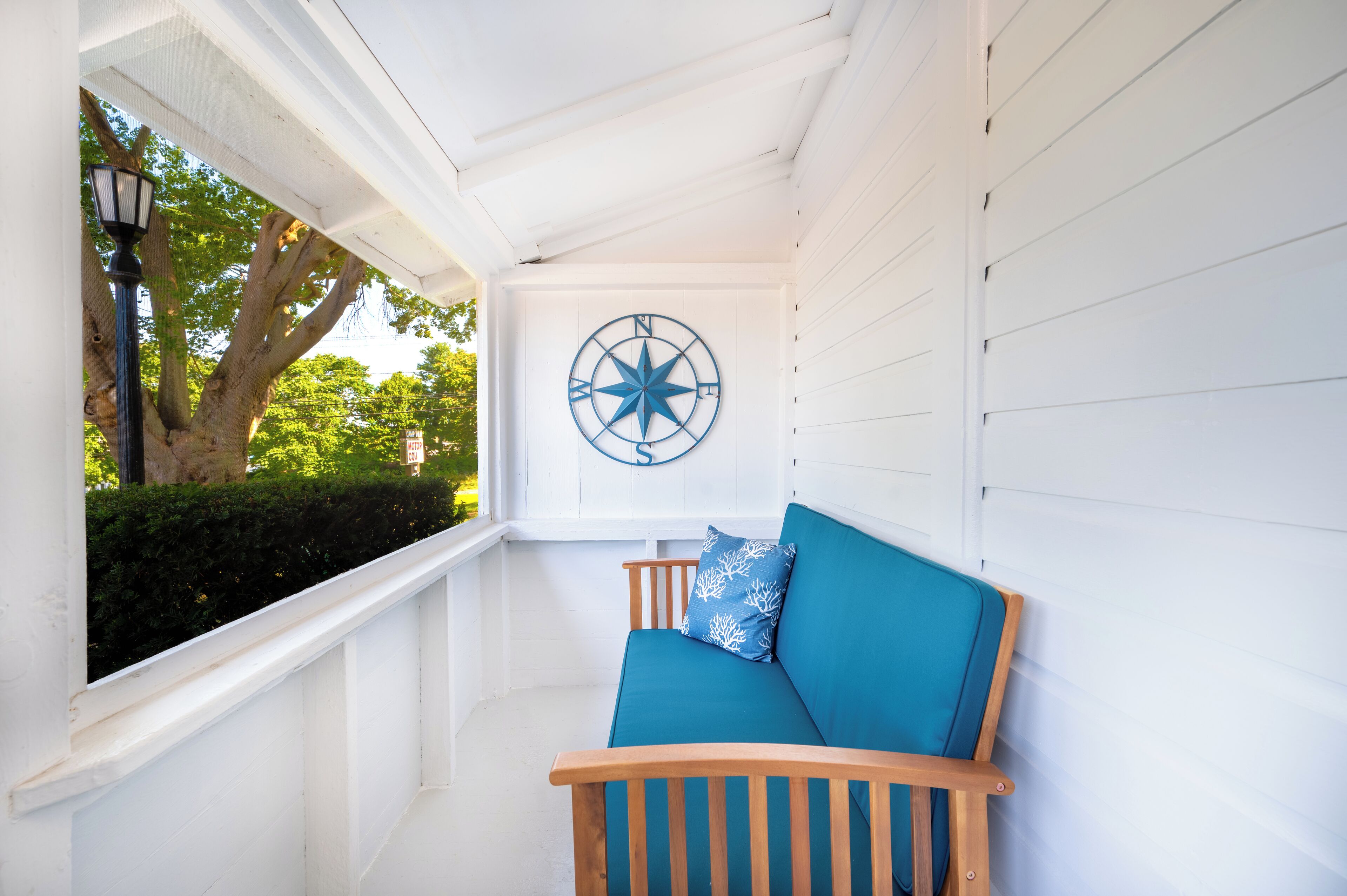 A white porch with a blue cushioned bench, a compass wall decoration, and a view of greenery.