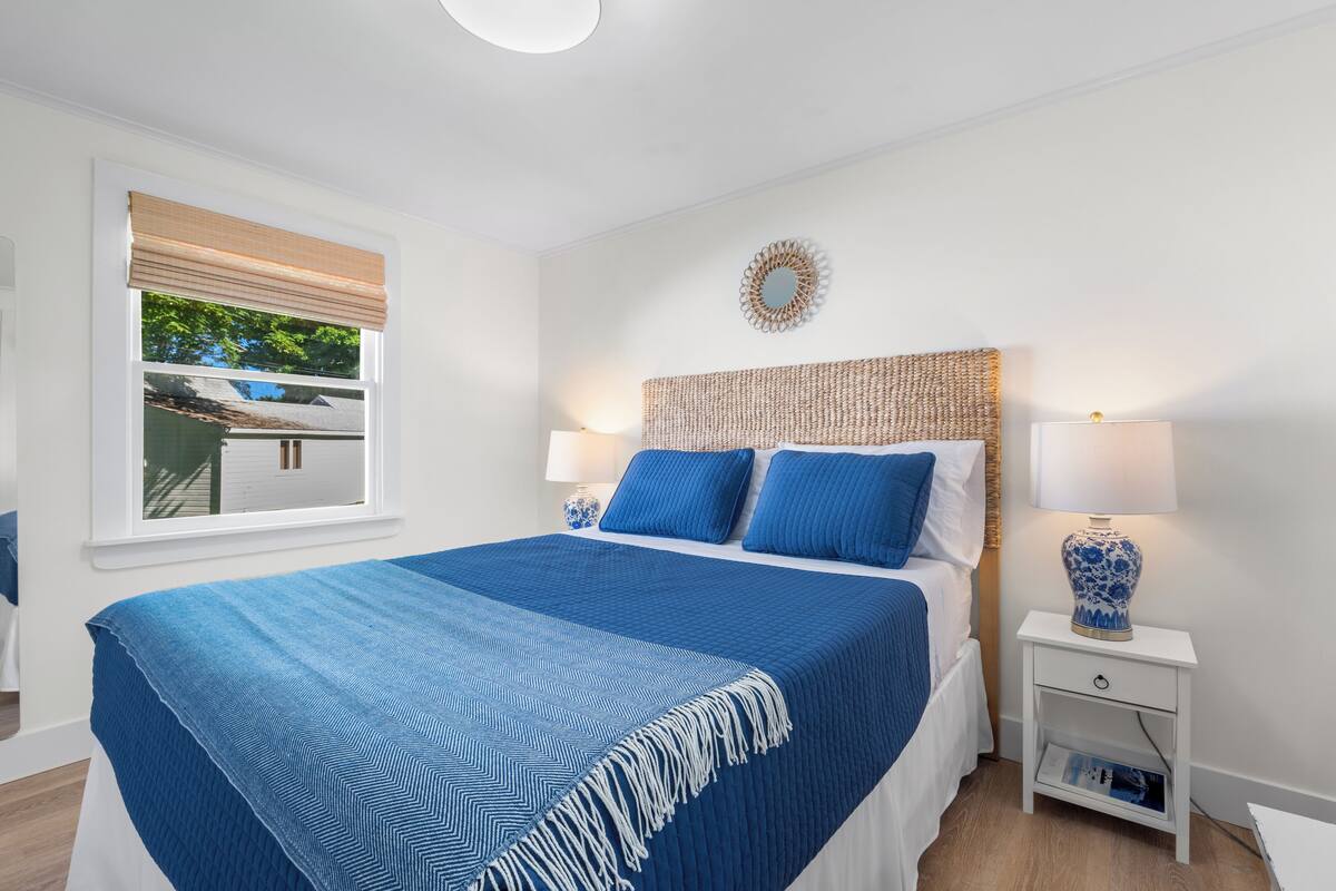 Une chambre à coucher avec un grand lit, deux tables de chevet, une fenêtre avec des stores et un miroir décoratif au mur.