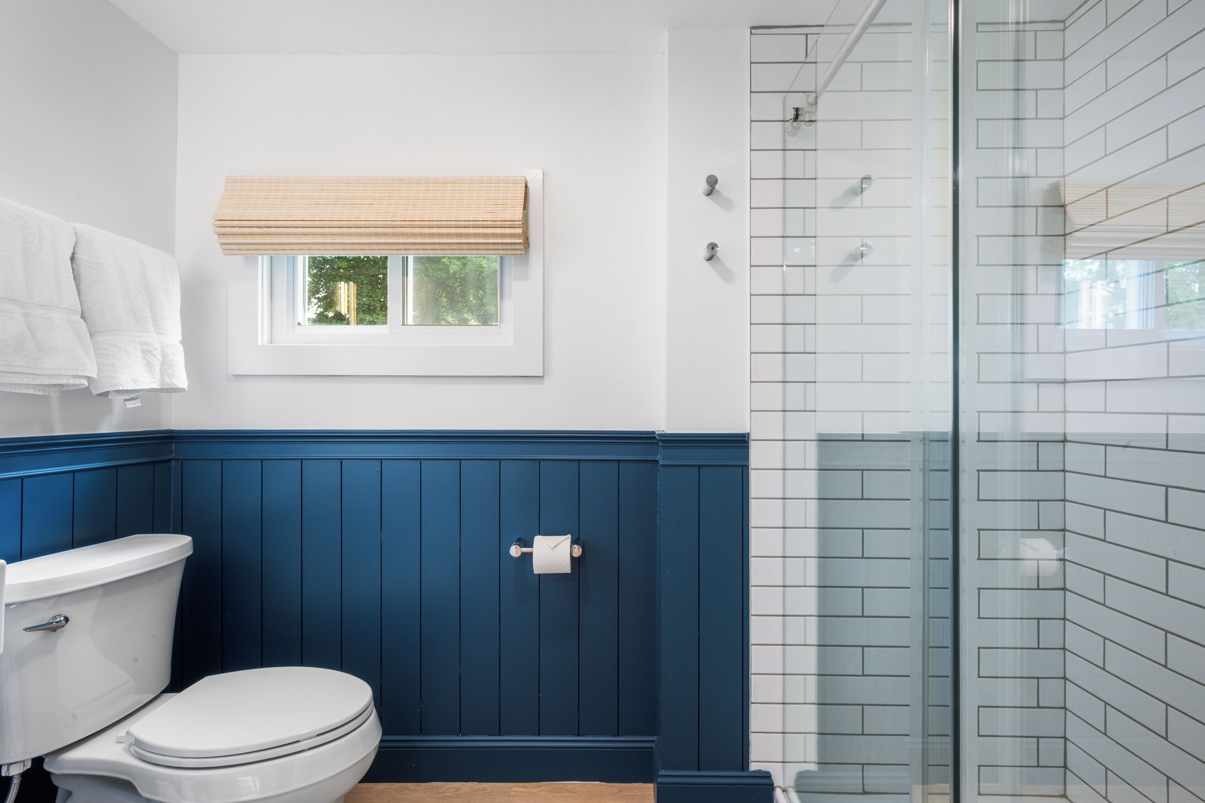 A bathroom with a white toilet, a blue wall, a window with a beige blind, and a glass shower enclosure.