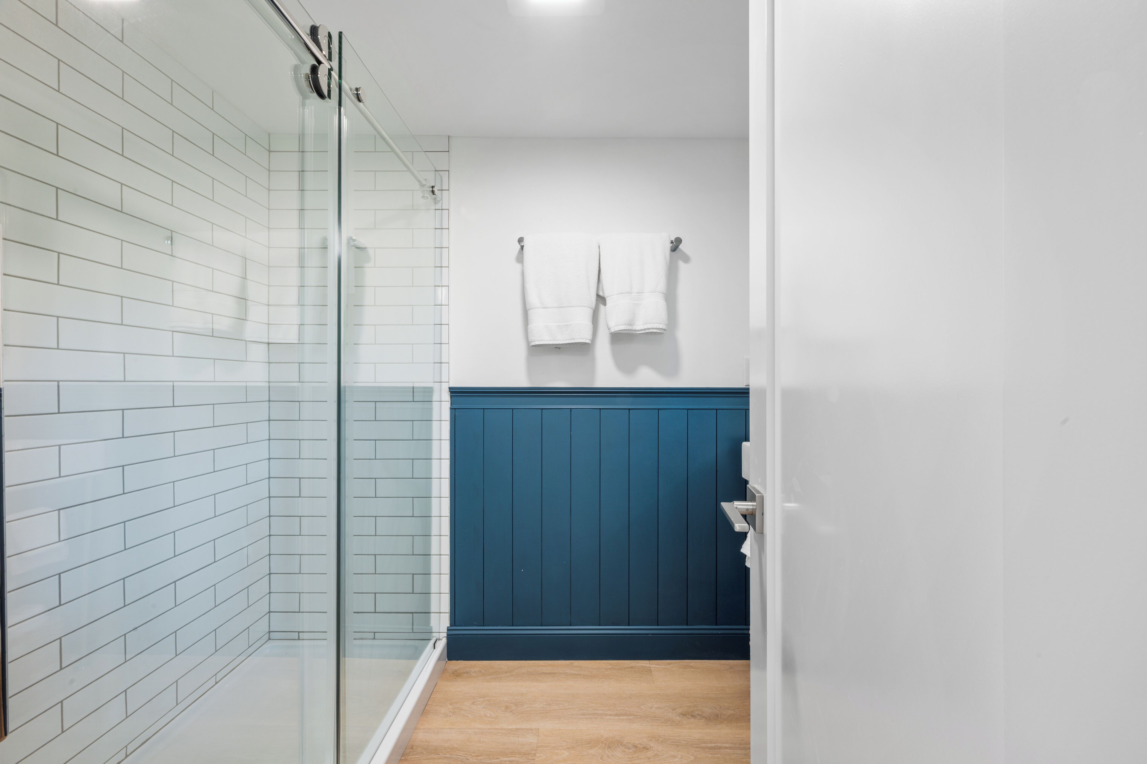 A bathroom with white tiled walls, a glass shower enclosure, a wooden floor, and a blue accent wall.