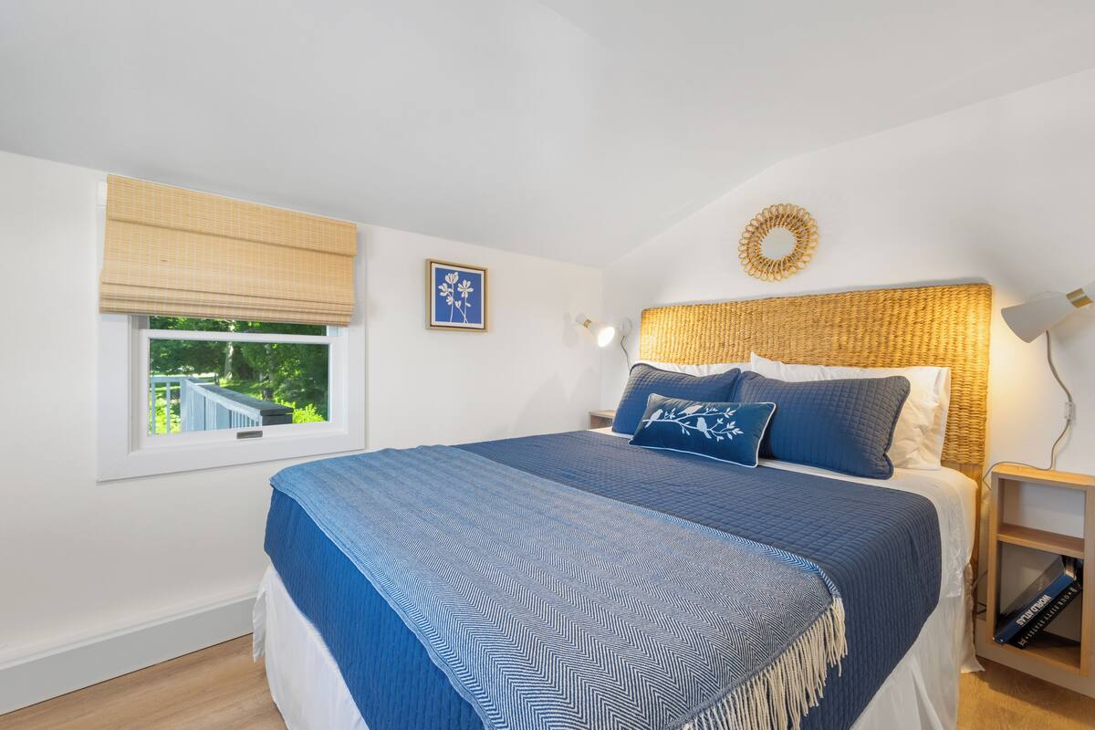 Une chambre avec un grand lit, une fenêtre avec des stores, une table de chevet avec une lampe et un élément de décoration murale.
