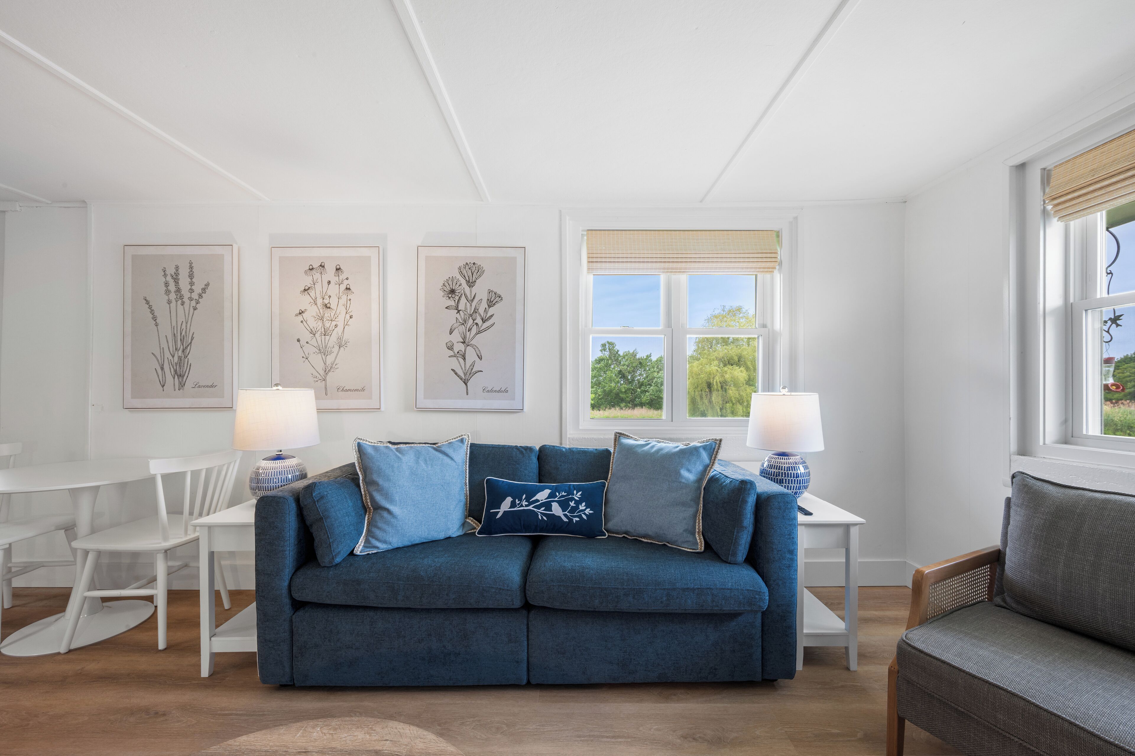 A living room with a blue sofa, white chairs, and framed botanical prints on the wall.