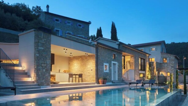 A modern villa with a swimming pool, stone exterior, and a well-lit interior.