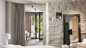 A modern bedroom with a large bed, a stone accent wall, a balcony with a table and chairs, and a view of greenery.