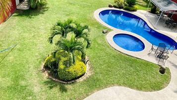 Un patio trasero con piscina, un porche con sillas, césped y arbustos bien cuidados.
