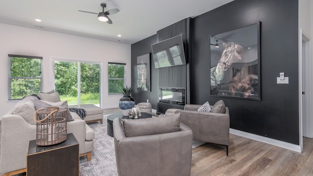 A modern living room with a grey sofa set, a coffee table, and a fireplace. There are large windows providing natural light and a view of greenery outside. - Newly Built Home w/ Fresh Design! (Columbia)