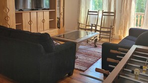 Ein Wohnzimmer mit einer Couch, einem Glastisch, einem Fernseher auf einer Holzkommode und einem Kicker.