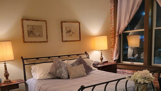 A bedroom with a bed, two bedside lamps, a nightstand, a chair, a glass table with a vase, and a window with curtains. - Downtown Cozy Castlerock Suite (Wenatchee)