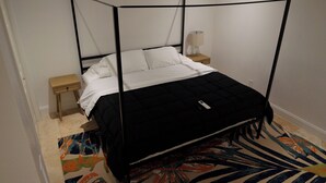 A single bed with a black quilted bedspread and white linens, a bedside table with a lamp, and a colorful abstract rug. - Bradenton Cozy Haven - Escape to our charming 3-bed, 2-bath Bradenton retreat! (Bradenton)
