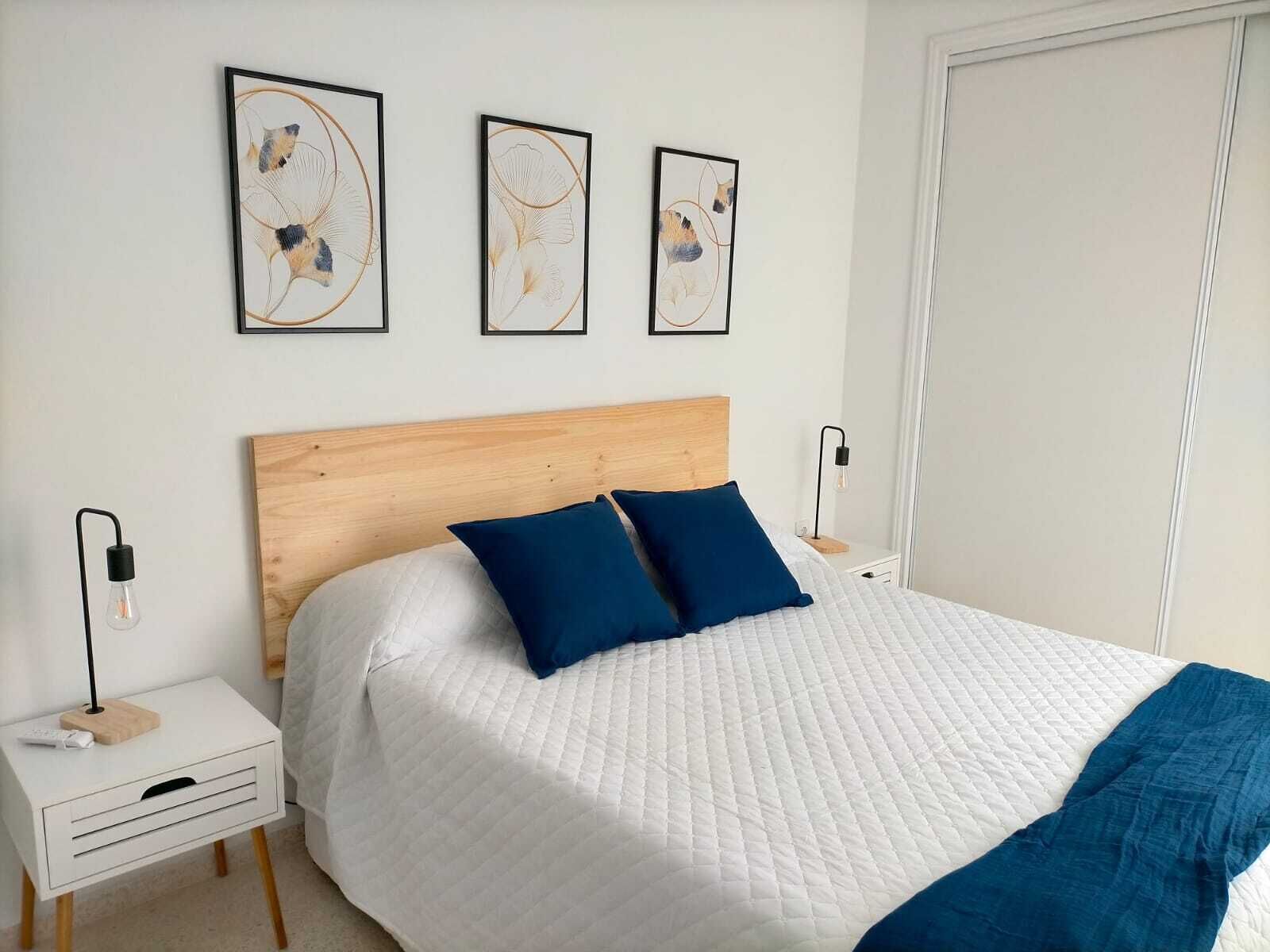 A neatly arranged bedroom with a wooden headboard, a white quilted bedspread, two blue pillows, and three framed artworks on the wall.
