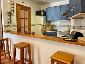 Una cocina con armarios blancos, un microondas, un refrigerador y una encimera de madera. Hay dos taburetes de madera y un jarrón con girasoles en la encimera.