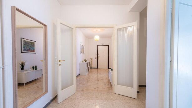 Un couloir aux murs blancs, doté d’un sol carrelé et d’une porte donnant sur une autre pièce.