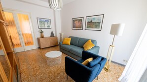 Un salon moderne avec un canapé gris, un fauteuil bleu et une table basse ronde. Deux œuvres d’art encadrées ornent le mur, et une lampe sur pied est placée dans un coin.