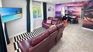 Uma sala de estar com sofá de couro, televisão, área de jantar com mesa e cadeiras, e um mural com o tema do pôr do sol na parede.