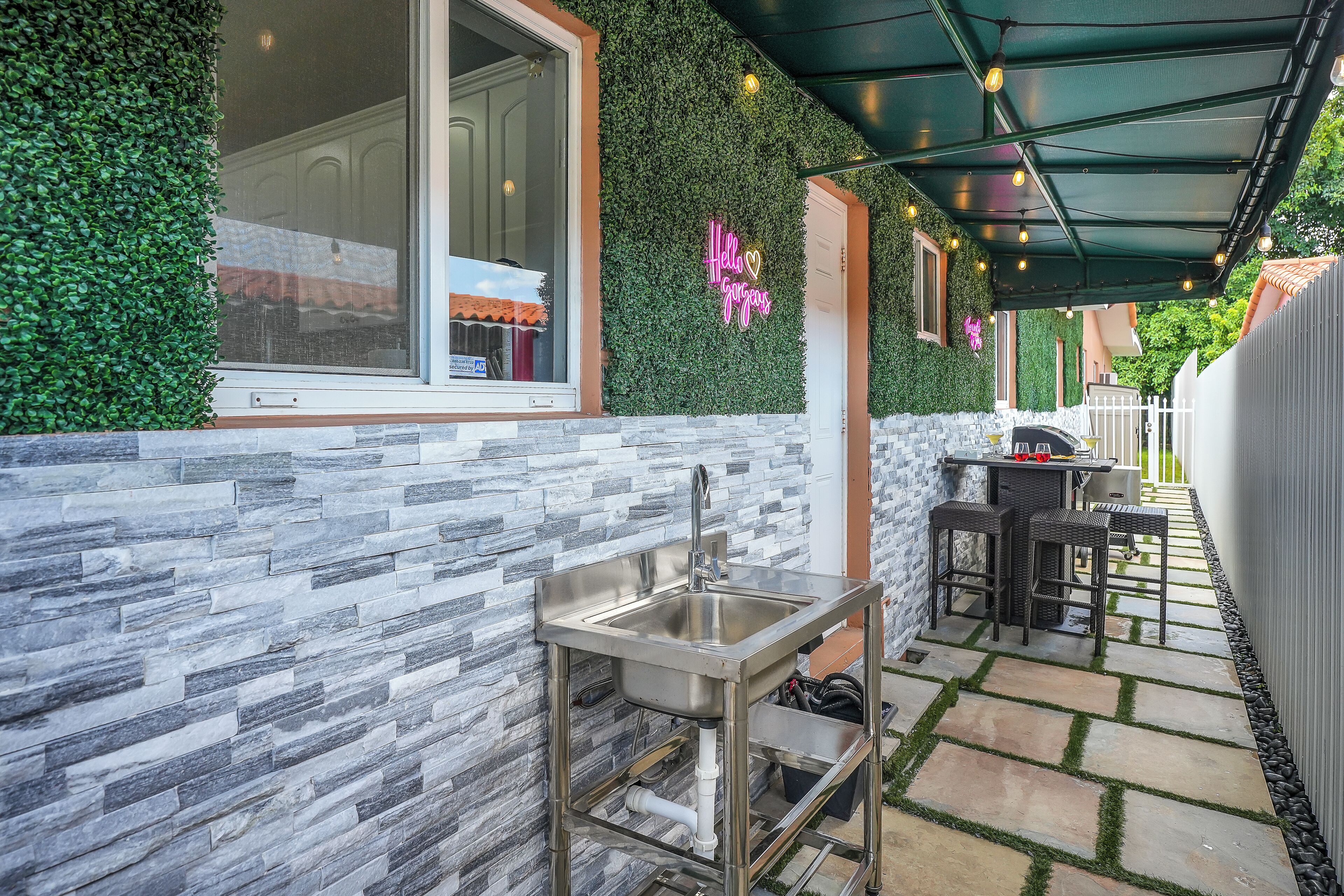 A modern outdoor kitchen area with a stone backsplash, a stainless steel sink, and a dining set under a green canopy.