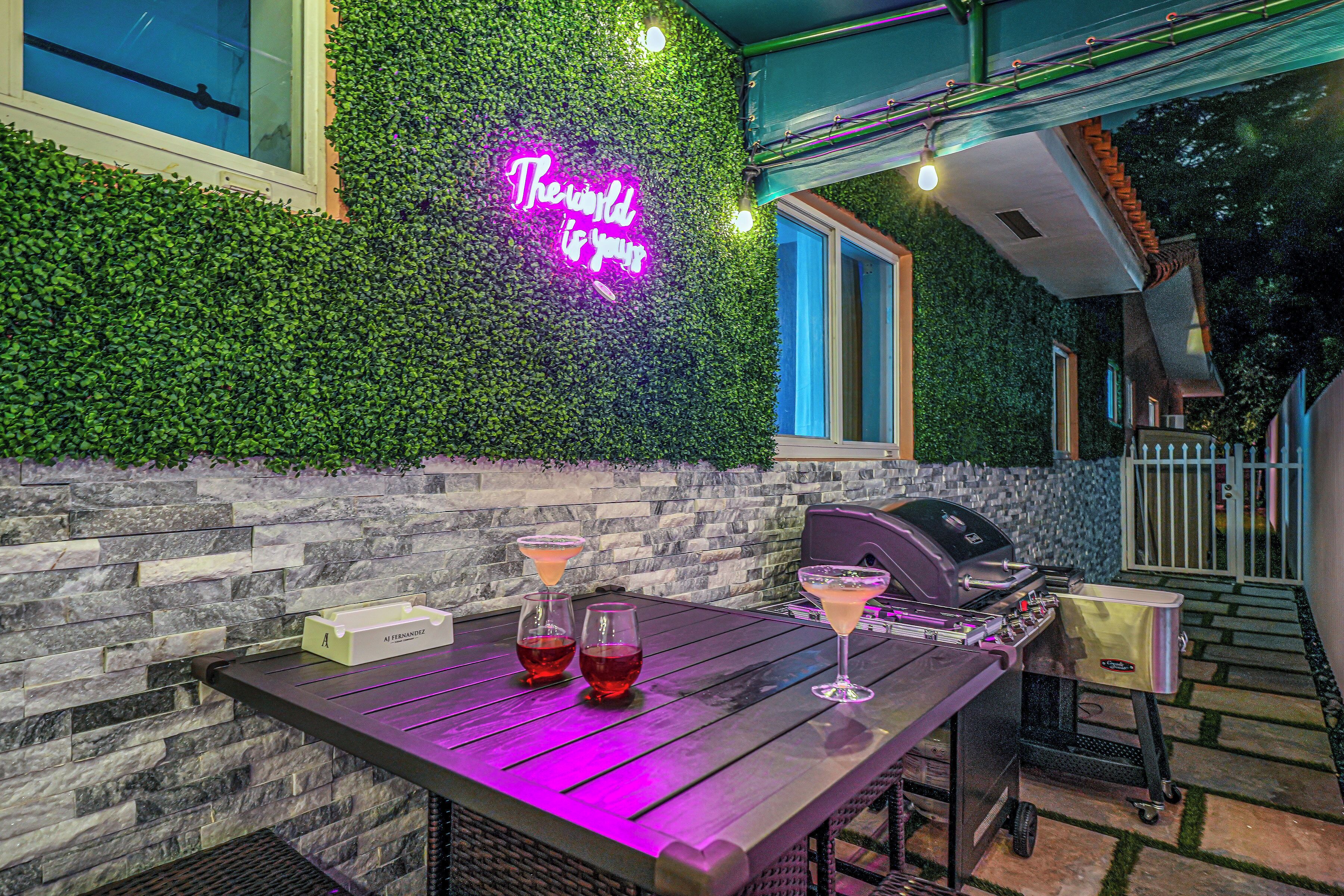 Un patio al aire libre con un letrero de neón, una parrilla y dos bebidas sobre una mesa.