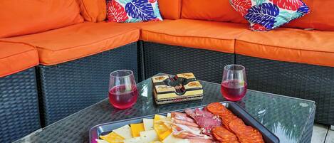 A glass-top table with a charcuterie board, two glasses of red drink, and a wicker sofa with orange cushions and colorful pillows.