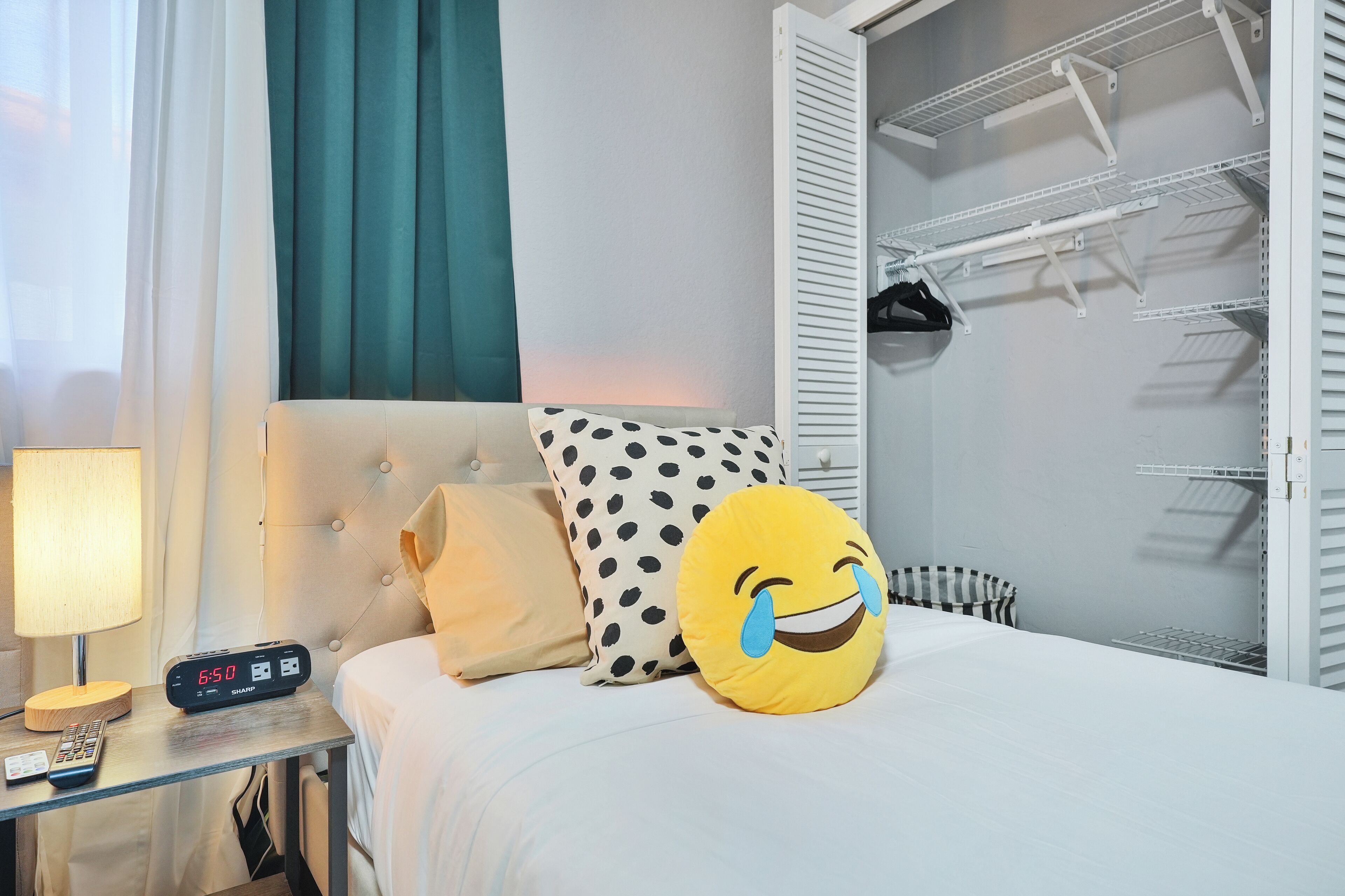 Una camera d'albergo con un letto, un comodino con una lampada e una sveglia digitale, un cuscino a pois e un cuscino a forma di emoji che ride, e un armadio con abiti appesi.