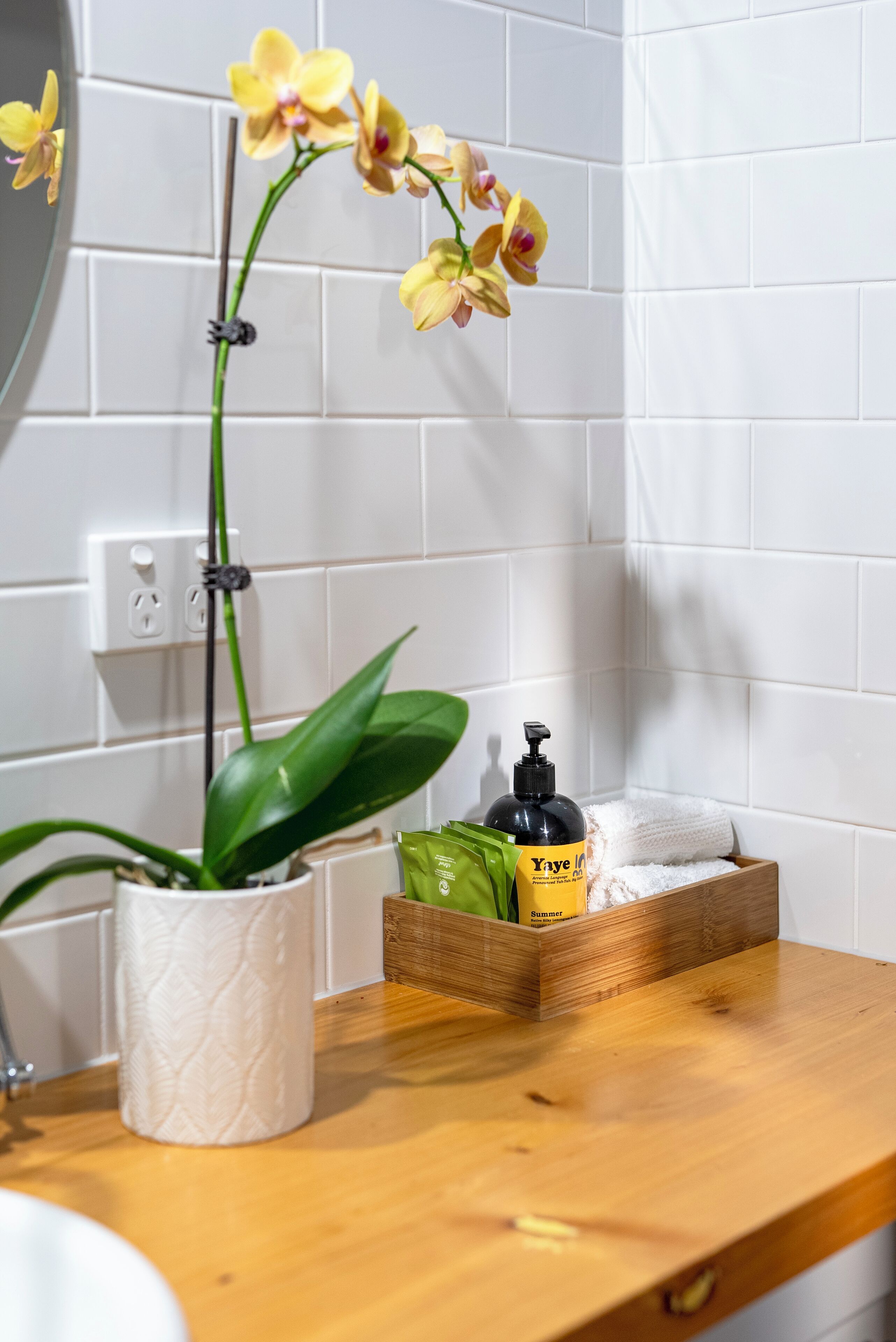 A bathroom with white tiles, a wooden counter, a potted orchid, a tissue roll, a green bag, and a bottle of shampoo.