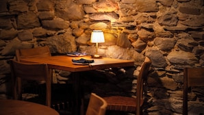Un mur en pierre, une table en bois, des chaises et une lampe de table dans une pièce à la lumière tamisée.