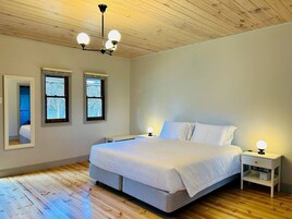 Quarto com cama, duas mesas de cabeceira, um criado-mudo, um espelho e teto de madeira.