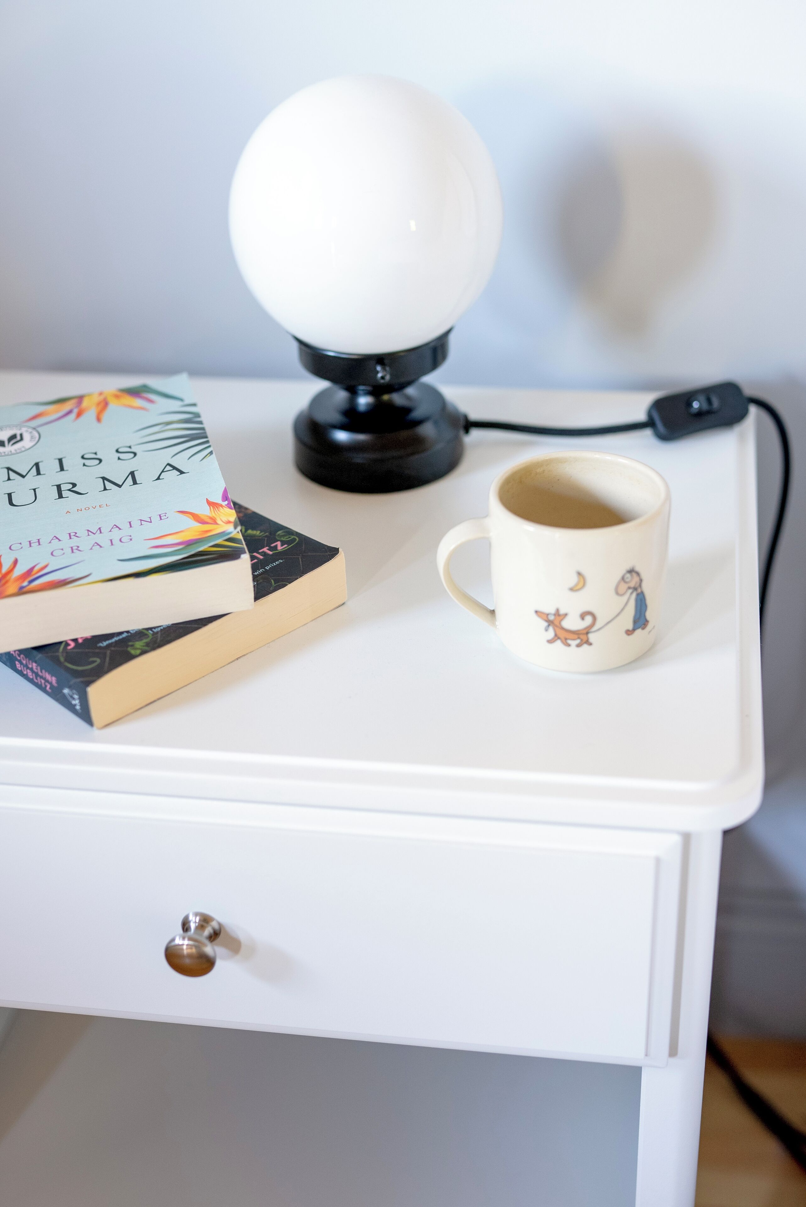 Ein weißer Nachttisch mit einer Lampe, einer Tasse und Büchern.