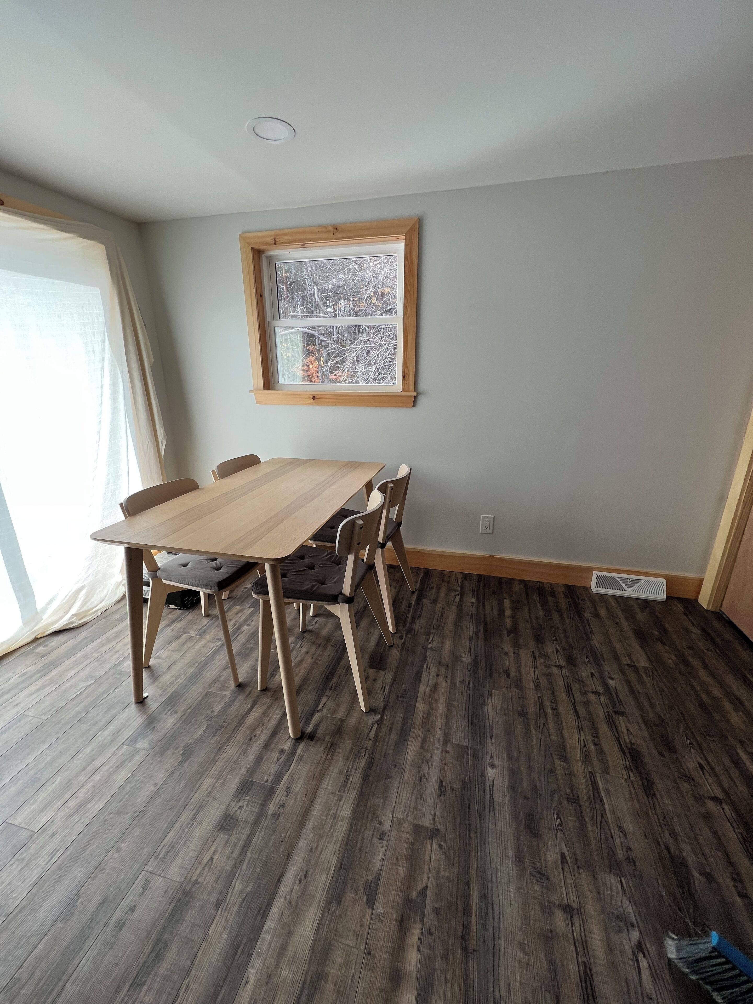 A dining area with a wooden table and chairs, a window with a view of trees, and a vent on the floor.